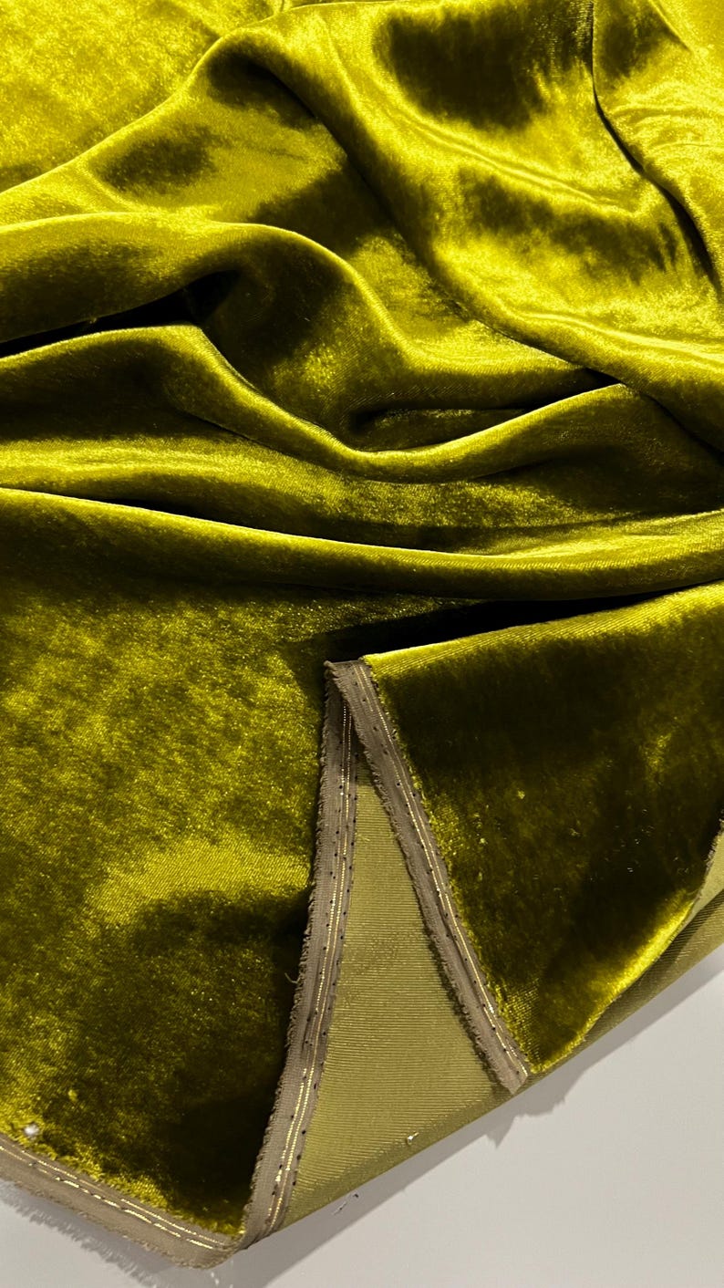 May include: A close-up of a rich, olive green velvet fabric with a subtle sheen. The fabric is folded and draped, showcasing its luxurious texture. The edges of the fabric are trimmed with a thin, gold-coloured thread.