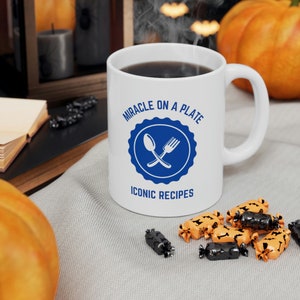 以下が含まれることがあります： 白いセラミック製のコーヒーカップで、青と白のデザインで「Miracle on a Plate Iconic Recipes」と書かれています。カップには、青い円の中に交差したスプーンとフォークがあります。カップは、オレンジと黒のキャンディーのある灰色の表面の上に置かれています。