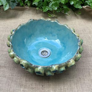 May include: A turquoise ceramic sink with a textured, scalloped rim in a contrasting green-yellow hue. The interior is a bright turquoise. The sink has a standard drain and is suitable for a bathroom or cloakroom.