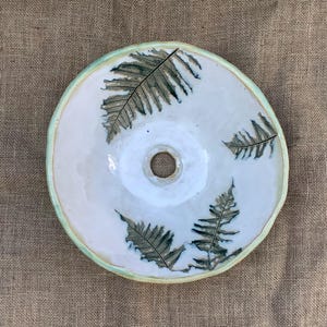 May include: A ceramic plate with a central hole, decorated with fern leaf patterns in green and brown on a white glazed surface. The plate has a light green rim. The plate is set against a neutral background.