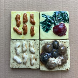 May include: Four square ceramic tiles with food designs. One tile features peanuts, another has a beet and leafy greens, a third shows shelled peanuts, and the last depicts a cluster of dark berries.