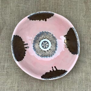 May include: A handcrafted ceramic plate with a pink base and dark brown detailing. The plate has a central decorative feature and a textured rim. The design is artistic, suitable for serving food or as a decorative item. The plate's diameter is approximately 25 cm.