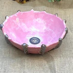 May include: A pink ceramic sink basin with a distinctive, scalloped edge design. The basin has a glossy finish and a drain in the centre. The edges are accented with dark grey details. This decorative sink is suitable for a bathroom.
