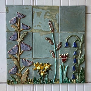 May include: Nine square ceramic tiles arranged in a 3x3 grid, featuring a floral design. The tiles have a muted teal background with raised relief flowers in shades of purple, yellow, and red. Green leaves and stems add contrast to the design.