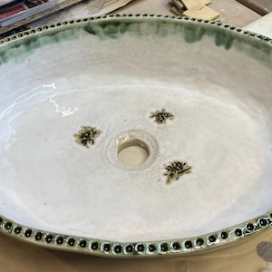 May include: Oval ceramic sink with a white interior and a green-glazed rim. The rim is decorated with small black dots. The sink features three decorative bee motifs and a central drain hole. A unique bathroom fixture.