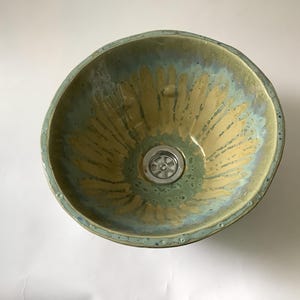 May include: A handcrafted ceramic sink basin with a unique, organic shape. The interior features a blend of green, blue, and gold hues, radiating from the drain. The rim has a textured, uneven edge, adding to its artisanal appeal.