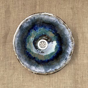 May include: A handcrafted ceramic sink with a unique, organic design. The sink features a circular shape with a dark blue and teal glazed interior, and a white drain cover. The rim has a textured, off-white finish.