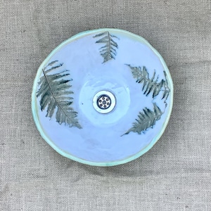 Könnte beinhalten: Ein handgefertigtes Keramikwaschbecken mit hellblauem Innenraum und hellgrünem Rand. Das Becken hat einen zentralen Abfluss und ist mit Farnblattmotiven in Grün- und Grautönen verziert.