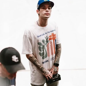 Könnte beinhalten: Ein junger Mann trägt ein weißes T-Shirt mit einem bunten Grafikdruck und dem Text "Speak Life, Be The Inspiration". Er trägt auch eine blaue Baseballkappe mit einem weißen Logo und eine silberne Kettenkette. Er hält eine schwarze Kamera in seiner rechten Hand.