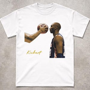 May include: White t-shirt with a basketball player and a basketball. The image includes the word "Kickart" in gold script. The basketball is held near the player's face.