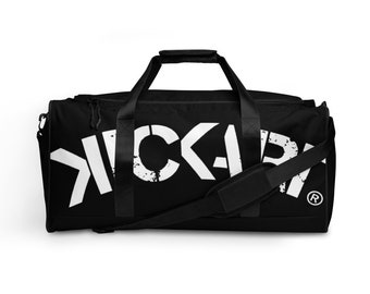 May include: Black duffel bag with white text that reads "KICKAP". The bag has a shoulder strap and two handles. The text has a distressed, grunge-like appearance. The bag is designed for carrying sports gear or travel items.