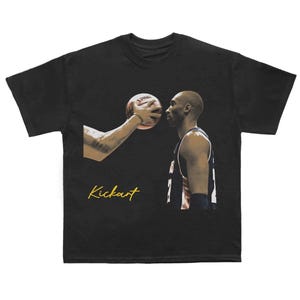 May include: Black t-shirt with a basketball player and a basketball, plus the gold script word "Kickart". The image shows a close-up of the player's face and a hand holding the basketball.