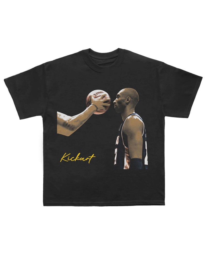 May include: Black t-shirt with a basketball player and a basketball, plus the gold script word "Kickart". The image shows a close-up of the player's face and a hand holding the basketball.