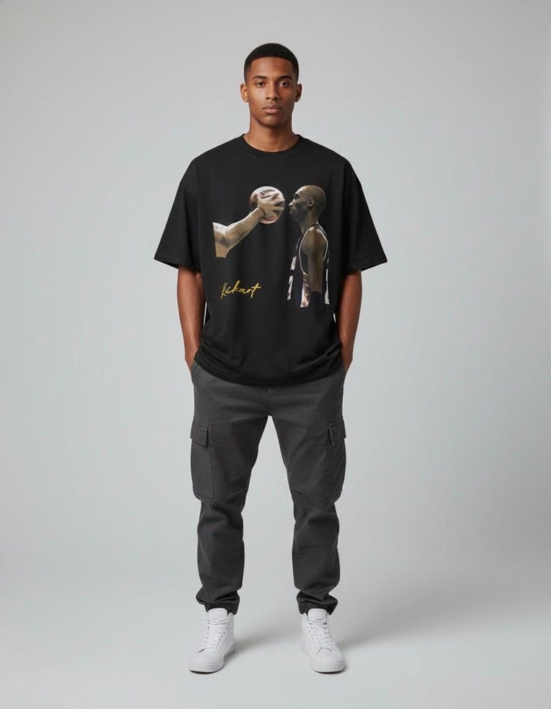 May include: Black t-shirt with a basketball graphic and the word "Kickart" in gold script. The person in the image is wearing dark grey cargo trousers and white trainers. The image depicts a basketball player and a basketball.