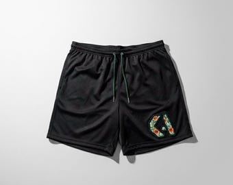 May include: Black athletic shorts with a drawstring waist and a floral "CA" patch. The shorts are made of a mesh material and have a green drawstring. The patch features red, green, and white floral accents.