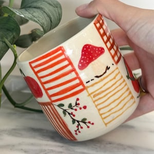 May include: A ceramic mug with a cream-colored base, adorned with hand-painted red and orange stripes, a red mushroom, and a sprig of berries. The mug is held by a hand, with a plant in the background.