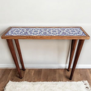May include: A wooden console table with a rectangular top featuring a blue and white floral tile design. The table has four angled, dark brown legs. The table is set against a white wall and a white rug.