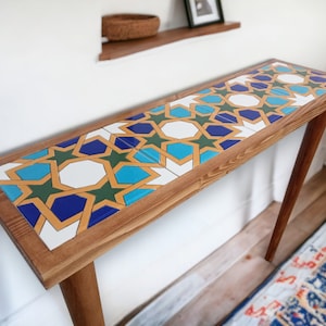 Puede incluir: Una consola de madera con una tapa rectangular con incrustaciones de azulejos estampados. Los azulejos presentan un diseño geométrico en tonos azules, verdes, blancos y dorados. La mesa tiene cuatro patas de madera en ángulo.