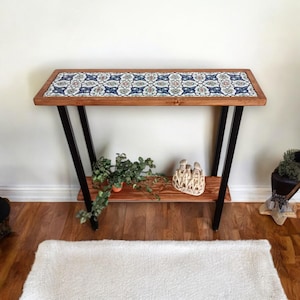 Könnte beinhalten: Eine Holzkonsolentisch mit einer gemusterten Fliesenoberfläche in Blau, Rot und Weiß. Der Tisch hat schwarze Metallbeine und ein unteres Regal. Eine kleine Topfpflanze und ein dekoratives Objekt befinden sich auf dem Regal.