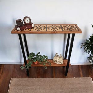 May include: A wooden console table with a geometric patterned top and a lower shelf. The table has black metal legs and is decorated with a small clock, a decorative box, and a potted plant.