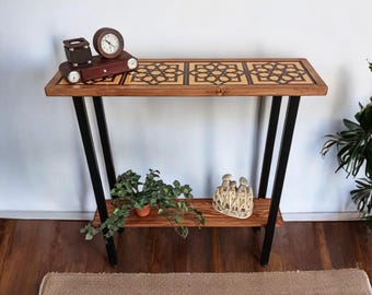 May include: A wooden console table with a geometric patterned top and a lower shelf. The table has black metal legs and is decorated with a small clock, a decorative box, and a potted plant.