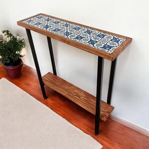May include: A rectangular console table with a wooden frame and a patterned tile top. The table has black metal legs and a lower shelf. The tile design features blue, red, and green floral patterns.