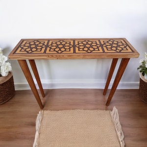 Puede incluir: Una consola de madera con una tapa rectangular con un patrón geométrico en marrón y beige. La mesa tiene cuatro patas anguladas y está sobre una alfombra marrón claro. La mesa mide aproximadamente 122 cm de largo.