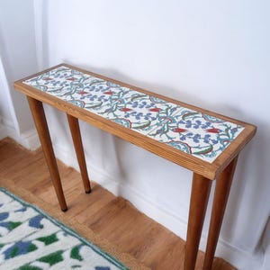Puede incluir: Una mesa consola larga y estrecha con un marco de madera y cuatro patas. La parte superior de la mesa está cubierta con azulejos blancos con un patrón floral azul y rojo.