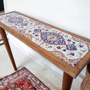 Può includere: Un tavolino da console in legno con un piano in piastrelle bianche con un motivo geometrico colorato. Il tavolo ha due gambe ed è alto circa 76 cm.