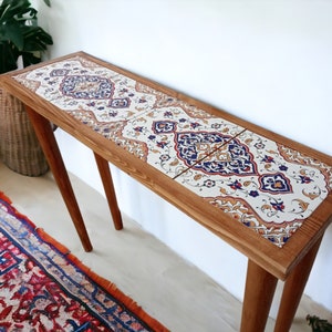 Può includere: Una consolle in legno con piano rettangolare intarsiato con piastrelle decorative. Le piastrelle presentano uno sfondo color crema con intricati motivi floreali blu, rossi e oro. Il tavolo ha quattro gambe in legno angolate.
