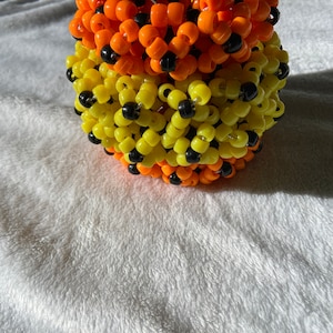 May include: A multi-colored kandi bracelet made of black, yellow, and orange beads. The bracelet is made in a cylindrical shape and is a popular accessory for raves and festivals.