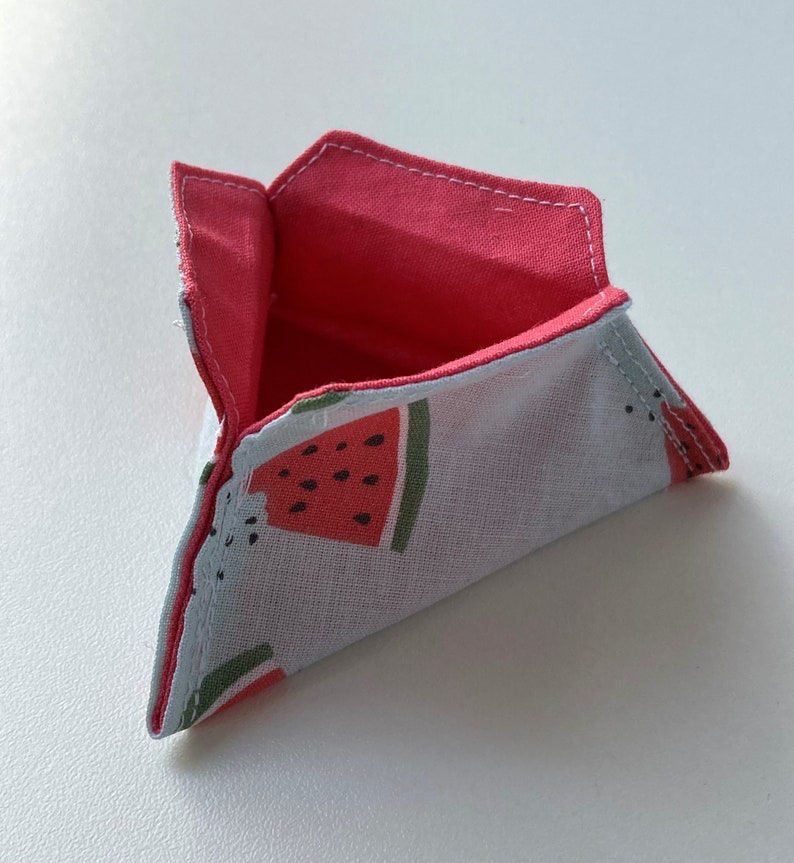 K&ouml;nnte beinhalten: Eine kleine, dreieckige Stofftasche mit Wassermelonenmuster. Die Tasche ist aufgeklappt und zeigt ein rosafarbenes Futter.