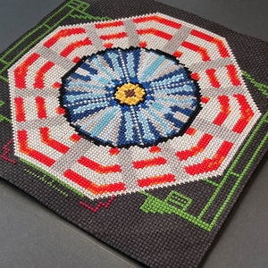 May include: A cross-stitch pattern of a colourful, geometric design with a blue, white and yellow centre. The design is surrounded by red and orange stripes.