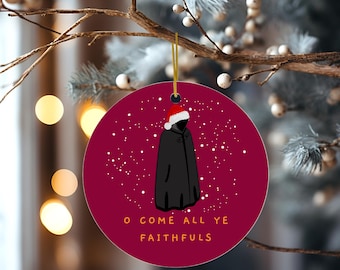 May include: A circular Christmas decoration with a burgundy backdrop. It displays a black figure in a Santa hat and cloak, with white snowflakes and the text "O COME ALL YE FAITHFULS". A gold string is attached for hanging on a Christmas tree.