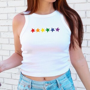May include: White ribbed tank top with a rainbow star design. The sleeveless top features a crew neck and a row of colorful stars across the chest. The top is paired with light blue denim jeans.