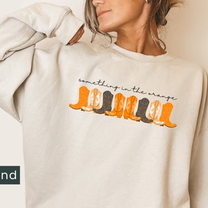 K&ouml;nnte beinhalten: Ein sandfarbener Pullover mit dem Text "something in the orange" und einer Reihe von orangefarbenen und schwarzen Cowboystiefeln, die auf der Vorderseite aufgedruckt sind.