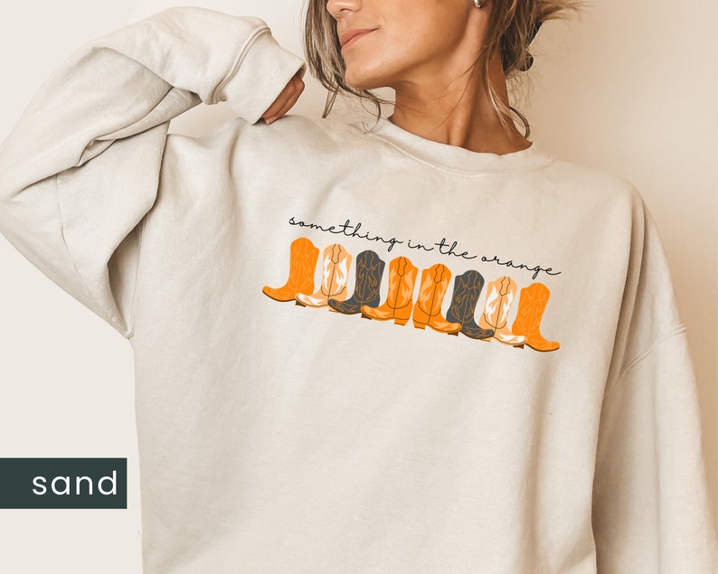 K&ouml;nnte beinhalten: Ein sandfarbener Pullover mit dem Text "something in the orange" und einer Reihe von orangefarbenen und schwarzen Cowboystiefeln, die auf der Vorderseite aufgedruckt sind.