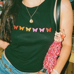 Puede incluir: Una camiseta sin mangas negra con una fila horizontal de siete pequeños gráficos de mariposas coloridas en tonos rojo, naranja, amarillo y rosa. La prenda tiene un cuello redondo acanalado y se combina con una falda de mezclilla azul y un cinturón de cadena dorado.