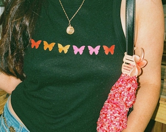 May include: A black sleeveless tank top featuring a horizontal row of seven small, colorful butterfly graphics in shades of red, orange, yellow, and pink. The top has a ribbed crew neckline and is worn with a blue denim skirt and a gold chain belt.