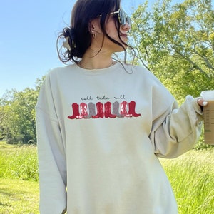 May include: A cream-colored sweatshirt with the text "roll tide roll" and a row of red and gray cowboy boots printed on the front.