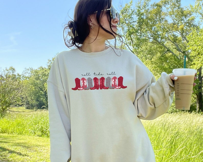 May include: A cream-colored sweatshirt with the text "roll tide roll" and a row of red and gray cowboy boots printed on the front.
