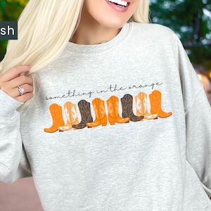 K&ouml;nnte beinhalten: Ein grauer Pullover mit dem Text "something in the orange" und einer Reihe von orangefarbenen und schwarzen Cowboystiefeln, die auf der Vorderseite aufgedruckt sind.