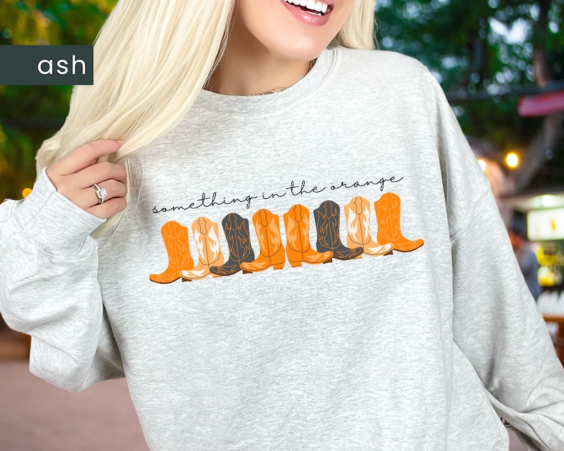 K&ouml;nnte beinhalten: Ein grauer Pullover mit dem Text "something in the orange" und einer Reihe von orangefarbenen und schwarzen Cowboystiefeln, die auf der Vorderseite aufgedruckt sind.