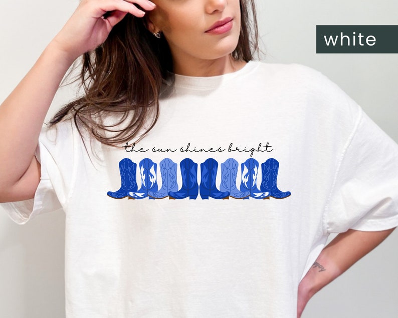 May include: White t-shirt with a blue graphic of cowboy boots and the text "the sun shines bright".
