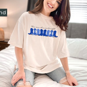 May include: A white t-shirt with a blue and white graphic of cowboy boots and the text "the sun shines bright".