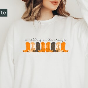 K&ouml;nnte beinhalten: Wei&szlig;er Pullover mit dem Text "something in the orange" und einer Reihe von orangefarbenen und schwarzen Cowboystiefeln, die auf der Vorderseite aufgedruckt sind.