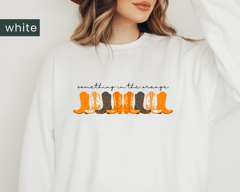 K&ouml;nnte beinhalten: Wei&szlig;er Pullover mit dem Text "something in the orange" und einer Reihe von orangefarbenen und schwarzen Cowboystiefeln, die auf der Vorderseite aufgedruckt sind.