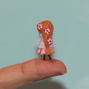 Può includere: Una piccola bambola all'uncinetto con lunghi capelli castani e fiori rosa nei capelli. Indossa un vestito rosa e bianco e sta in piedi su un dito.