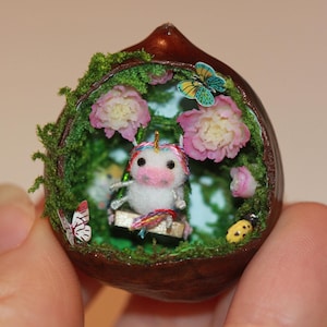 May include: A small, white unicorn figurine with a pink nose and rainbow mane sits on a swing inside a hollowed-out chestnut shell. The shell is decorated with moss, pink flowers, and butterflies.