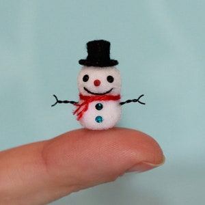 Könnte beinhalten: Ein winziger weißer Schneemann mit einem schwarzen Hut, einem roten Schal und blauen Augen. Der Schneemann sitzt auf einem Finger.